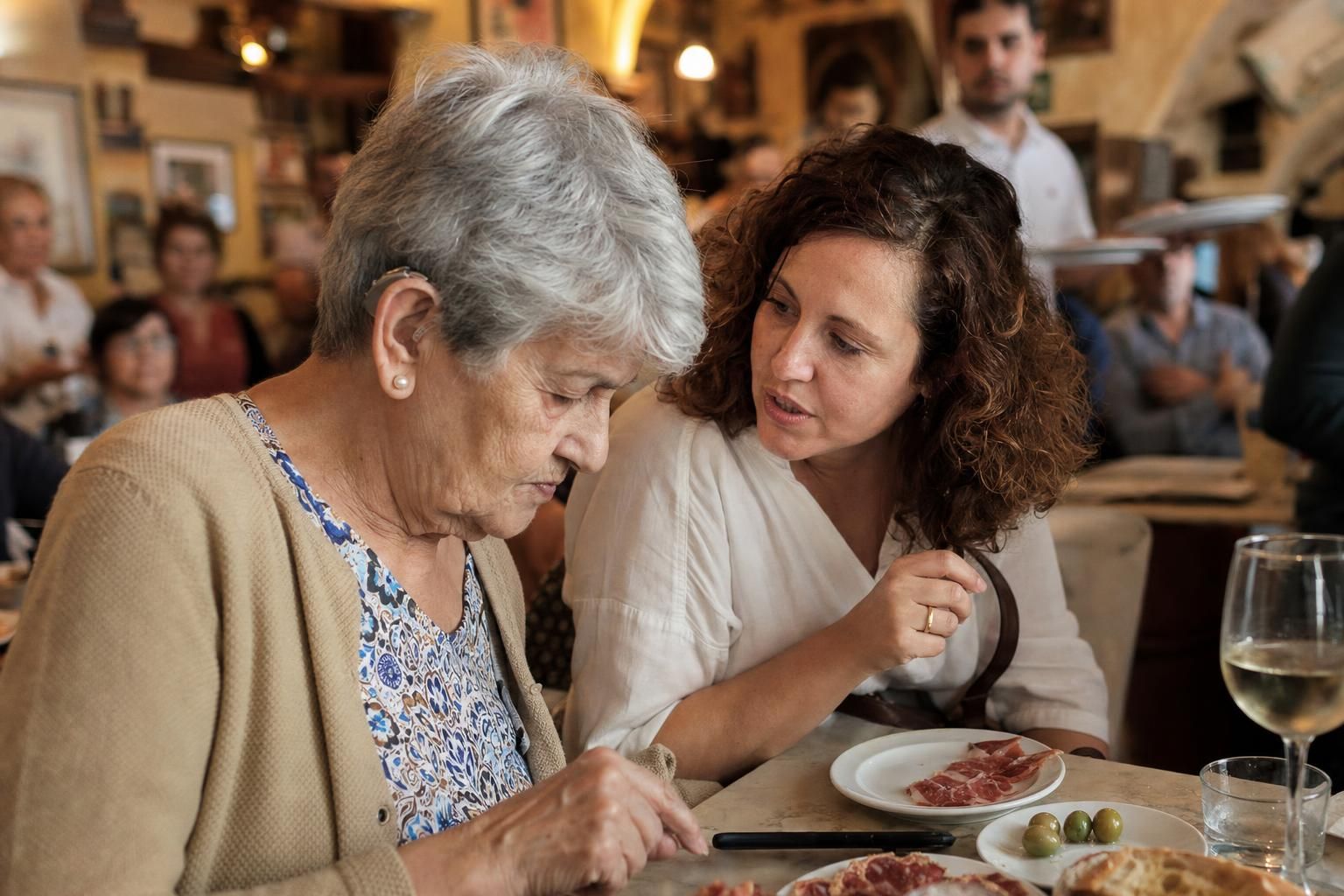 In a busy restaurant, a woman with a hearing aid reads her phone on the table while her daughter talks to her.